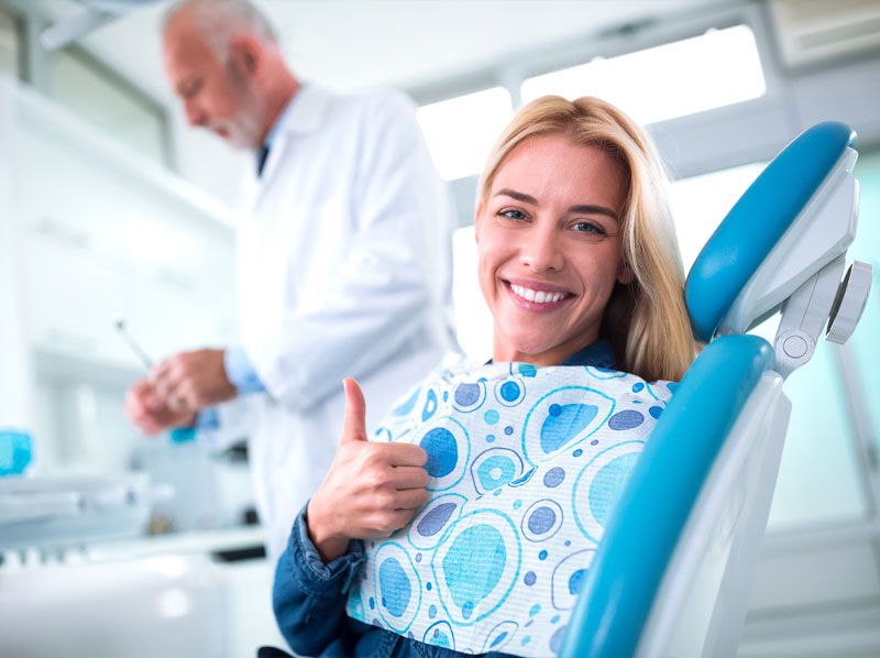 Dental patient sitting in dental chair giving a thumbs-up while doctor is in the backgound working.